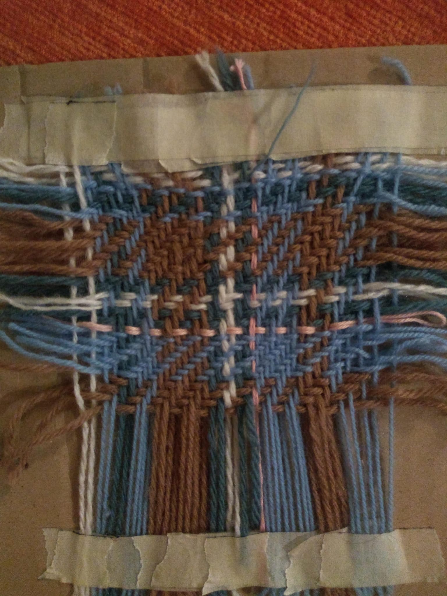A small tartan sample woven on a cardboard loom, in blue, rust, and dusty pink threads