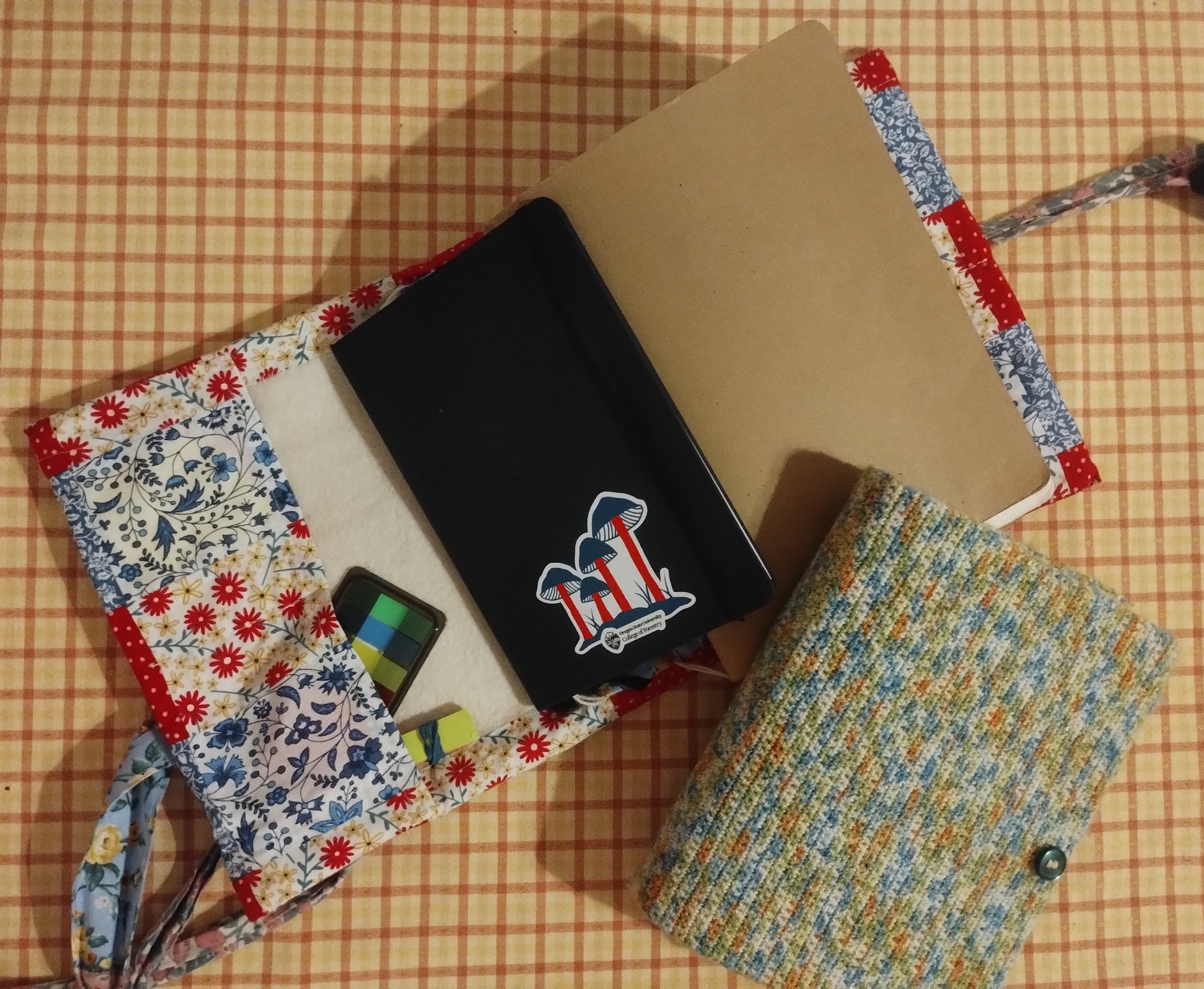 A fabric-covered notebook, a crocheted book cover, and a tablet laid out on a checked tablecloth