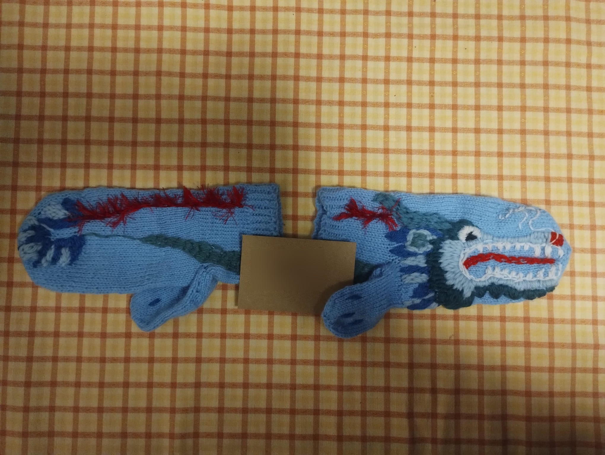 A pair of pale blue hand-knitted mittens embroidered with a dragon, its body and teeth stretching across both hands
