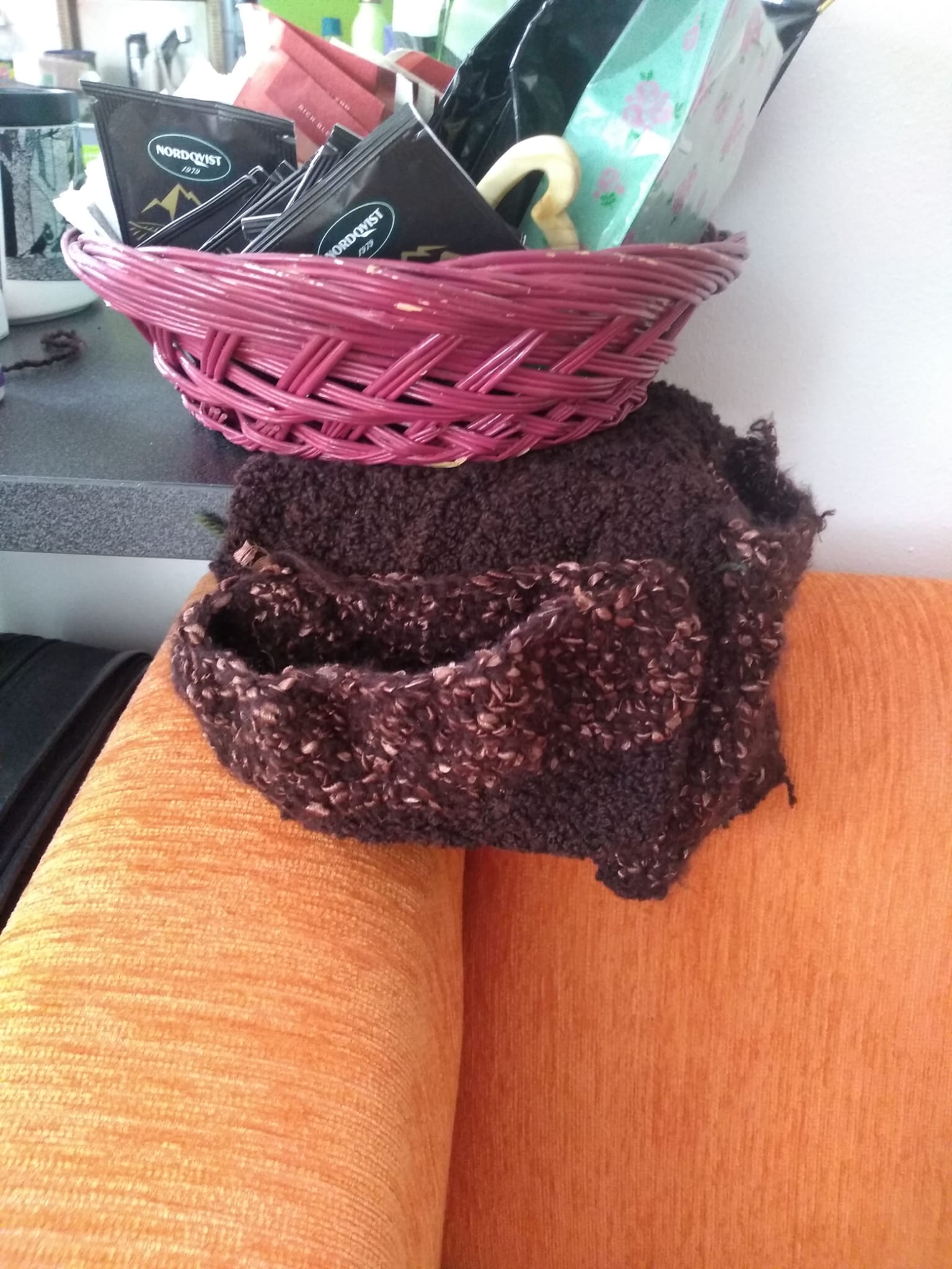 A small knitted brown pouch with pockets, perched on the arm of an orange sofa