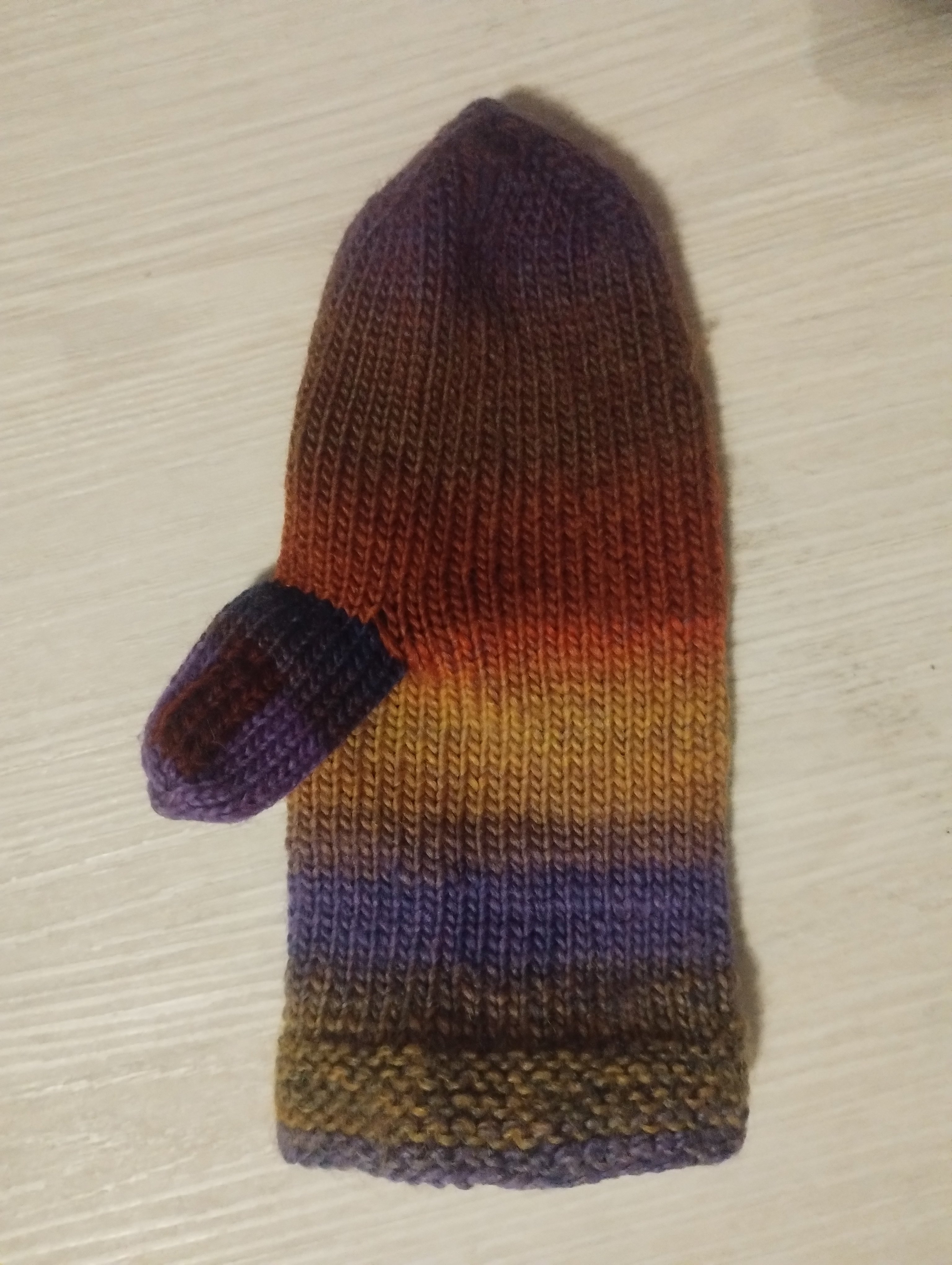 A hand-knitted mitten in a rainbow gradient of purple, gold, and rust