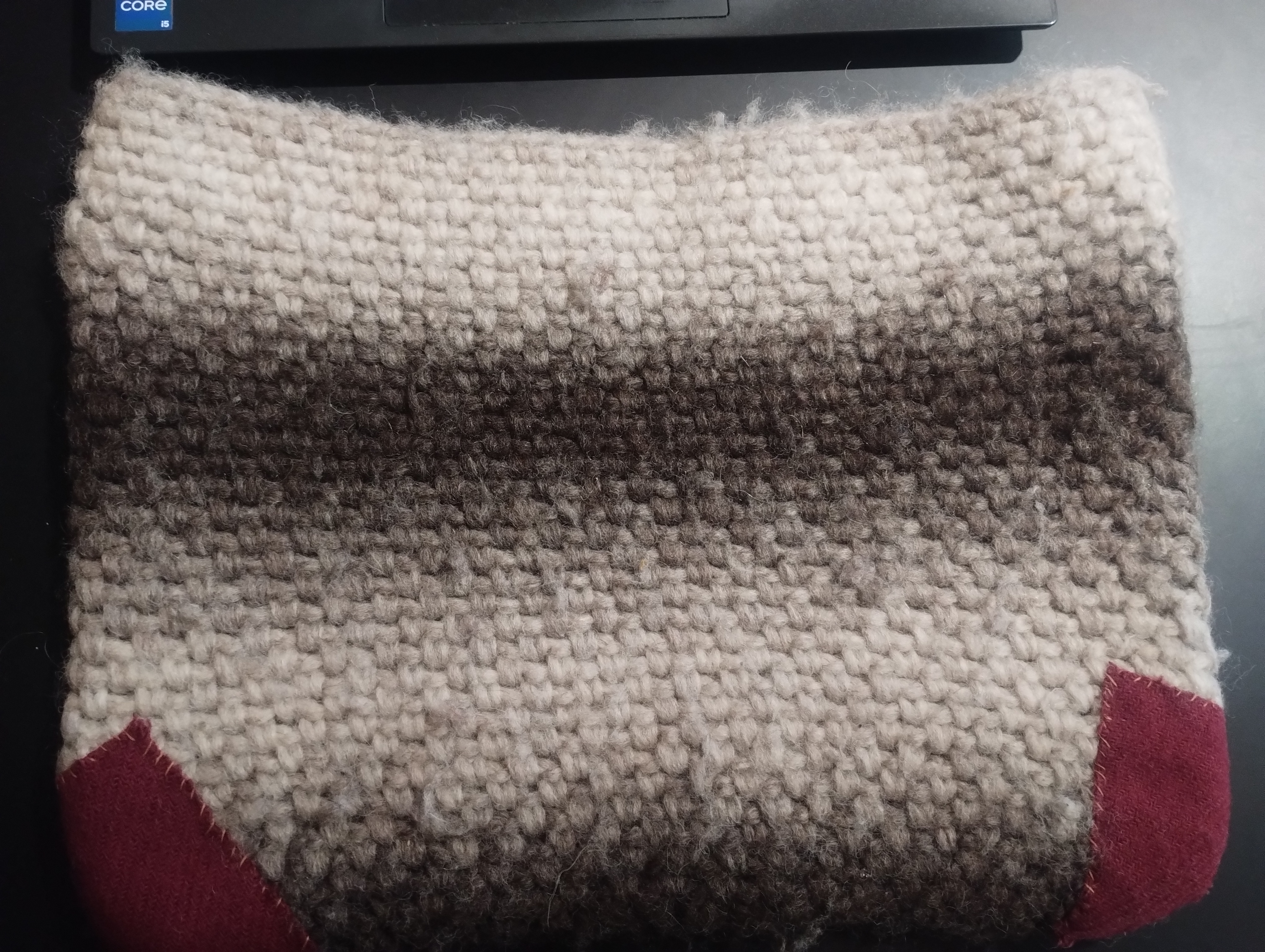 A beige and brown crocheted laptop sleeve with mended burgundy wool patches stitched onto both bottom corners