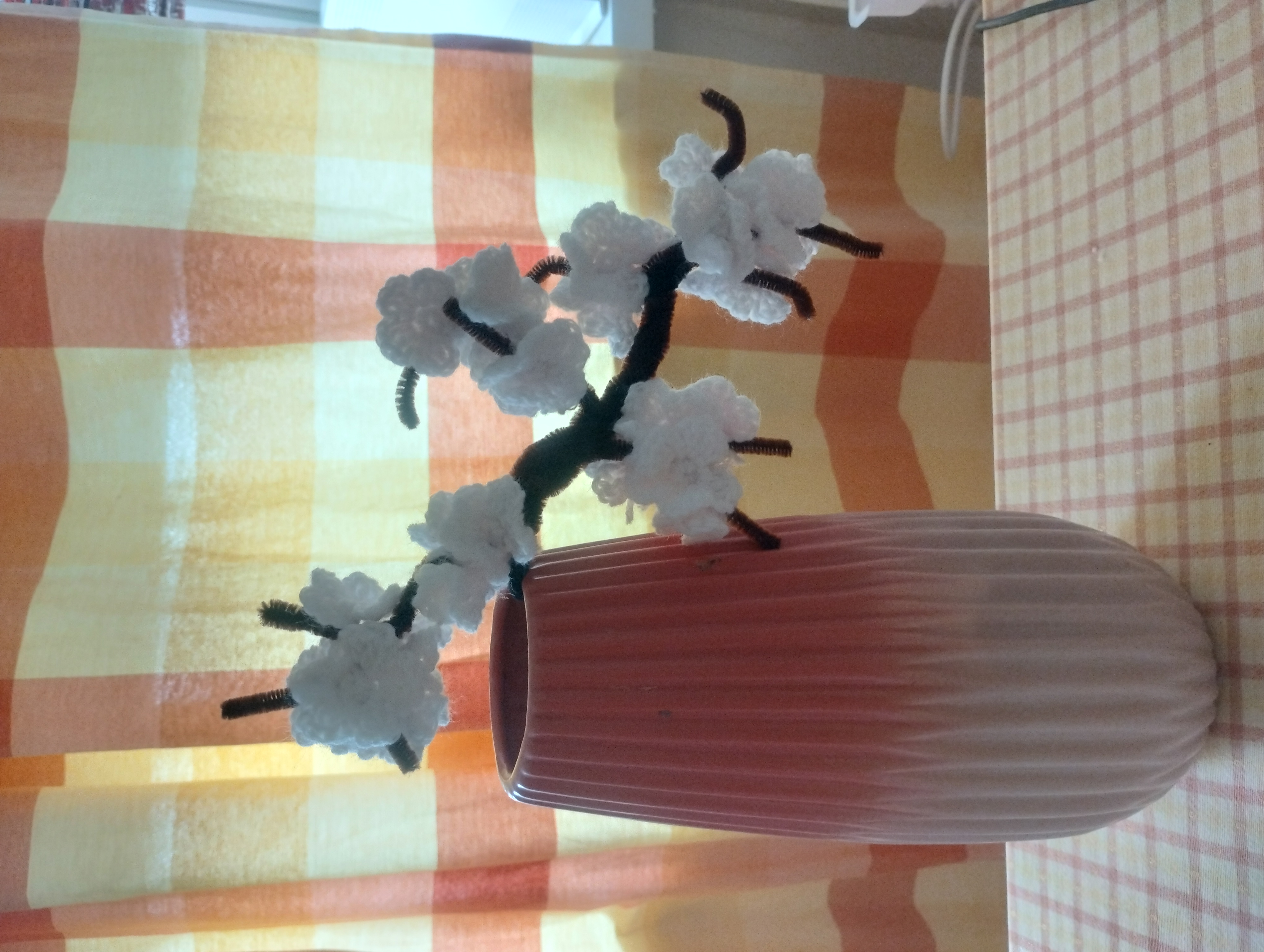 A crocheted blossom branch with white flowers on wire stems, arranged in a pink vase