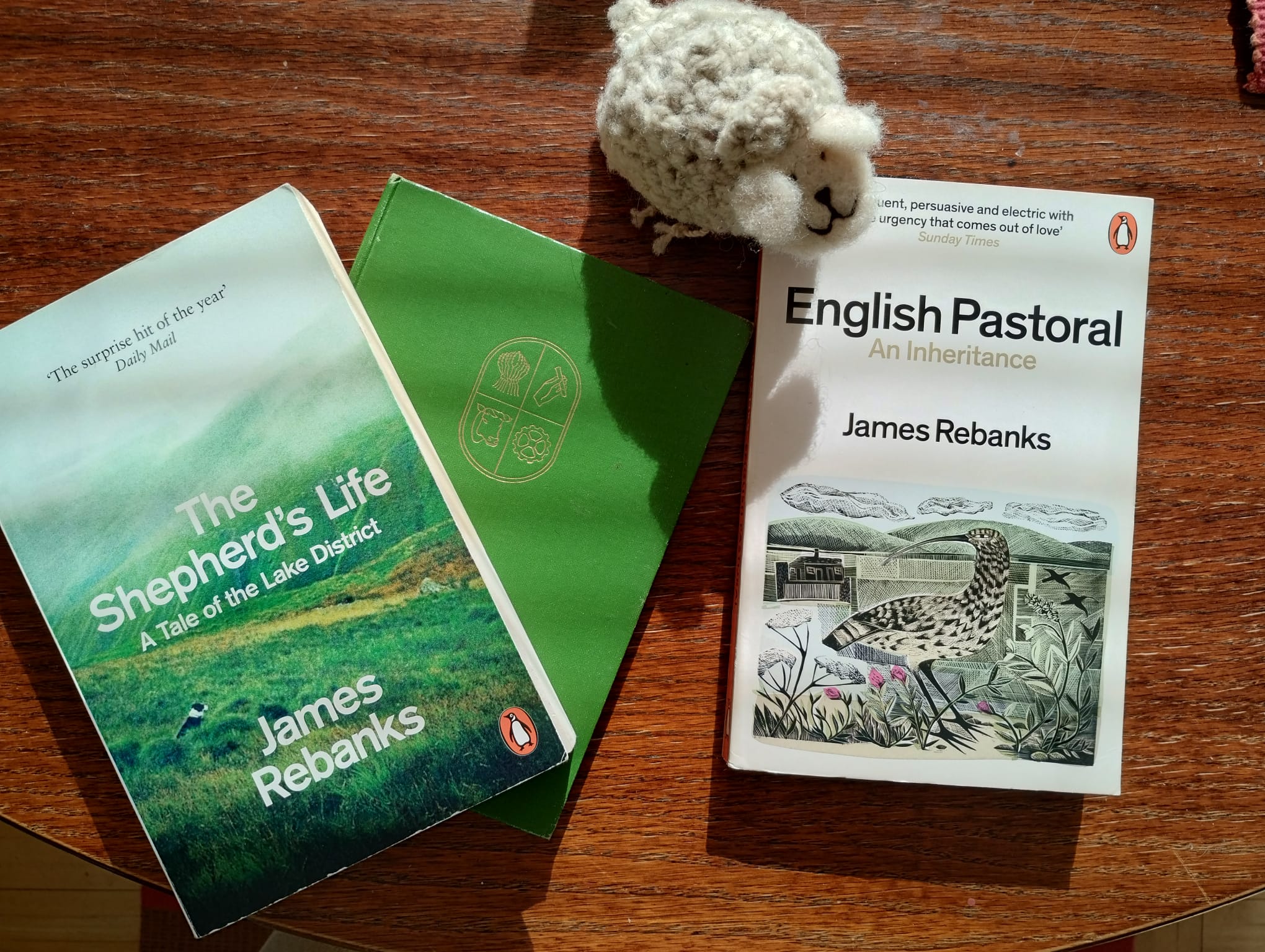 A small needle-felted sheep sitting on top of two James Rebanks books about sheep farming