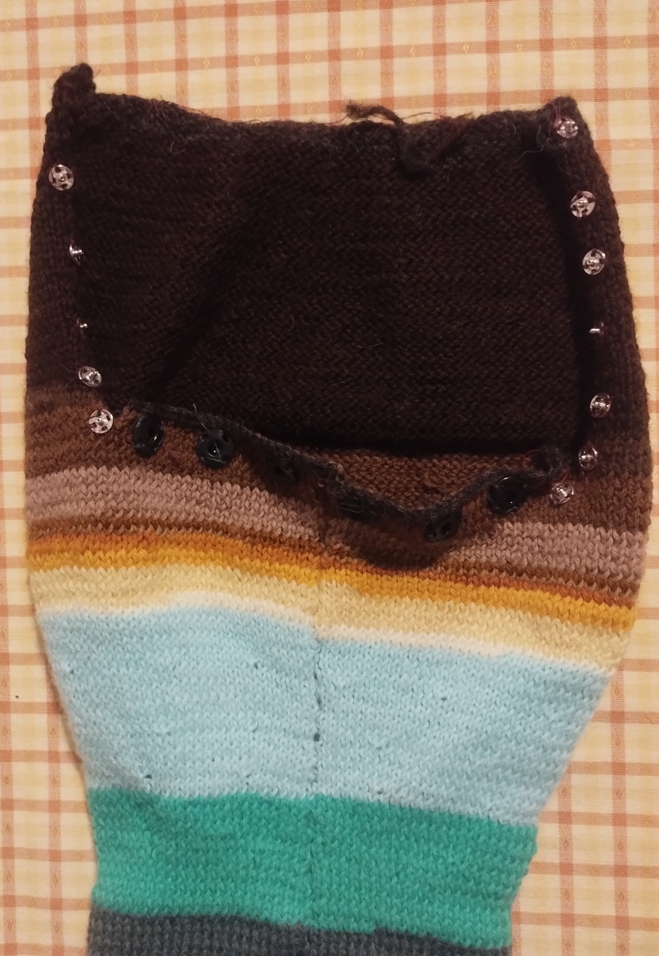 A striped knitted garment in brown, mustard, teal, and green, with a row of buttonholes down one edge