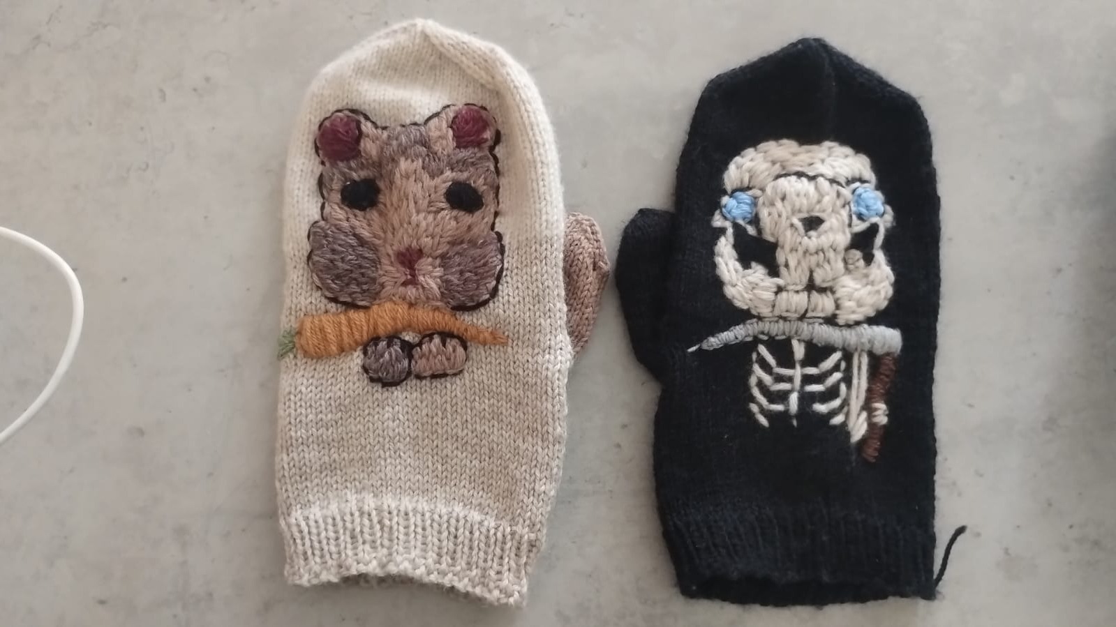 A pair of hand-knitted mittens, one with a hamster holding a carrot and one with a skeletal Death figure carrying a scythe