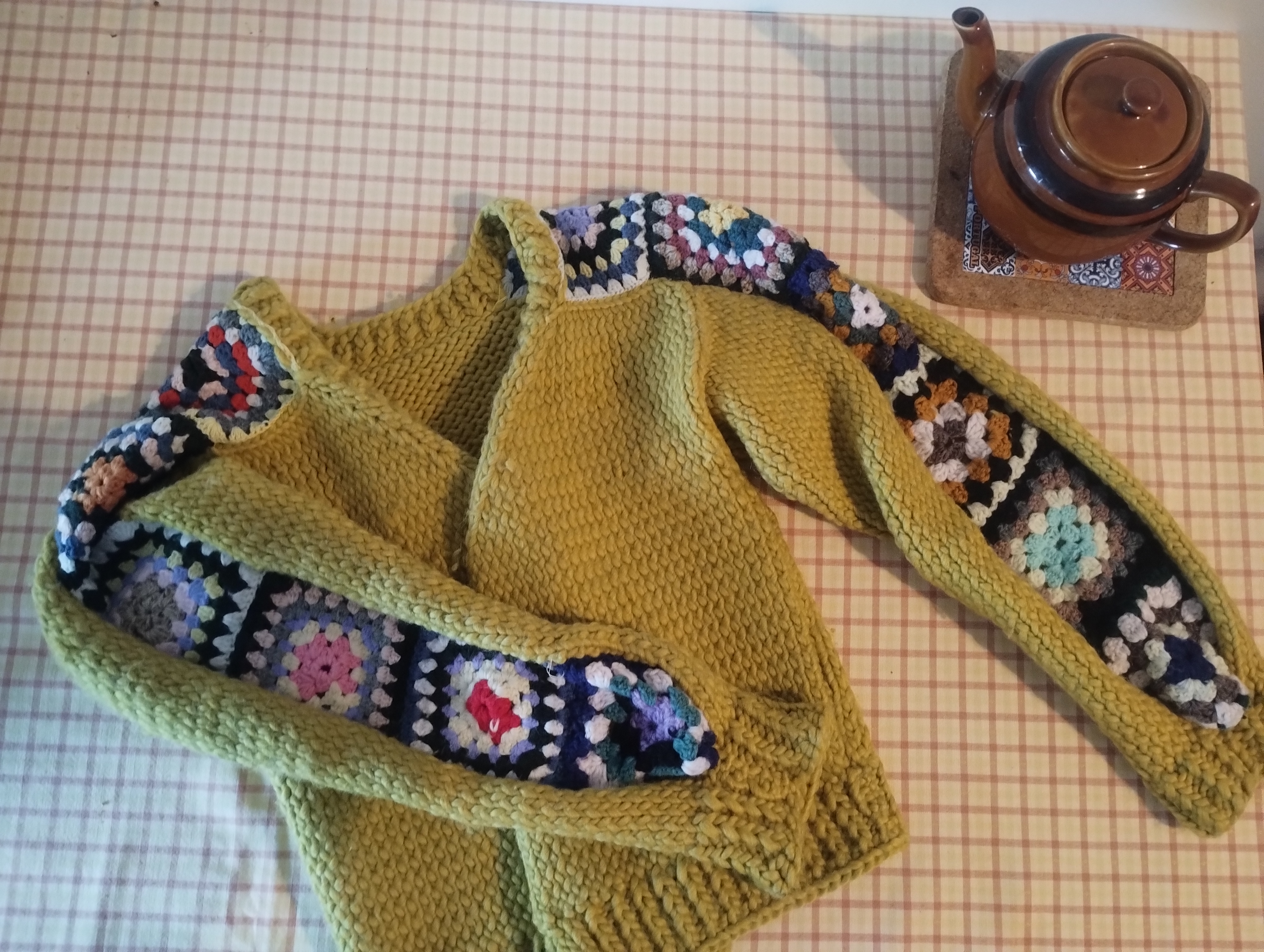 A mustard-yellow knitted cardigan trimmed with colourful granny squares along the front edges and collar